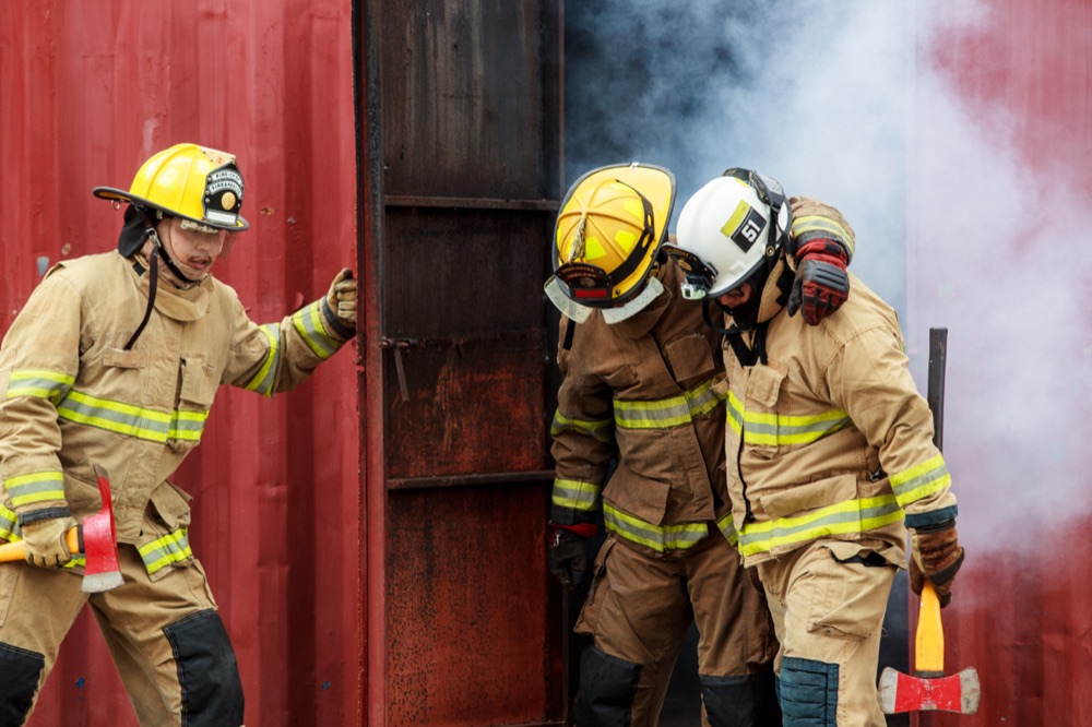 Two firefighters in full gear supporting each other while emerging from smoke.